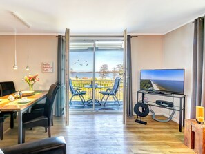 A modern living room with a dining area, a TV on a stand, and a balcony with a view of the sea. - Residenz am Balmer See, apartment 61 with sauna, lake view and wellness (Balm)