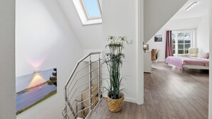 Un intérieur moderne avec un escalier, une grande plante en pot, un tableau représentant un coucher de soleil et une chambre à coucher avec un lit et des rideaux.