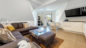 A modern living room with a sofa, ottoman, and coffee table, a flat-screen TV, and a kitchen area with cabinets and a countertop.