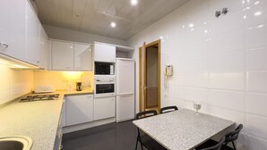 Una cocina compacta con armarios blancos, microondas, horno y lavaplatos. Hay una pequeña mesa de comedor con sillas.