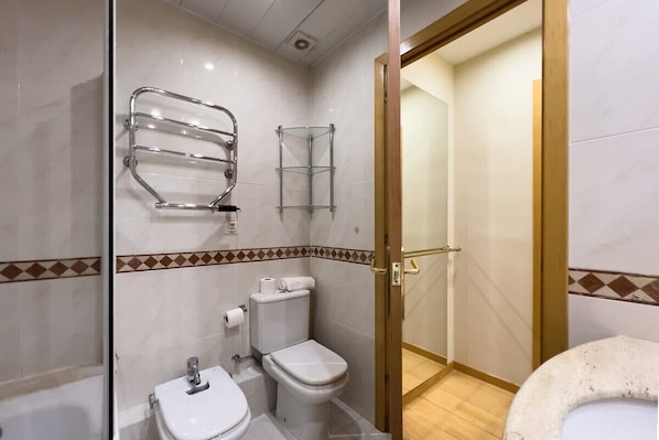 A bathroom with a toilet, sink, and shower area. There are towel racks on the wall and a toilet paper holder. The walls have a patterned tile design. - Gran Via Sun (Barcelona)