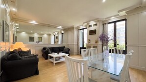 Amplia sala de estar con una mesa de comedor de vidrio, sillas blancas, un sofá negro y una mesa de centro blanca.