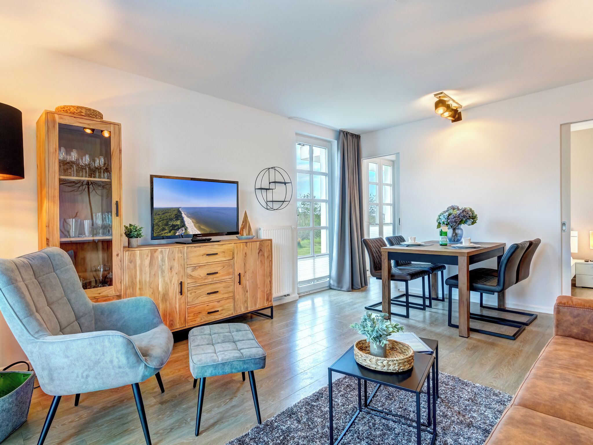 A modern living room with a flat-screen TV, wooden cabinet, grey armchair, dining table with chairs, and a small side table with a plant.
