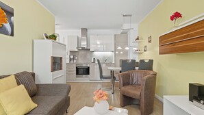 Un salon moderne avec un canapé, un fauteuil et une table basse. On aperçoit, au fond, une cuisine avec des meubles blancs et des appareils électroménagers en acier inoxydable.