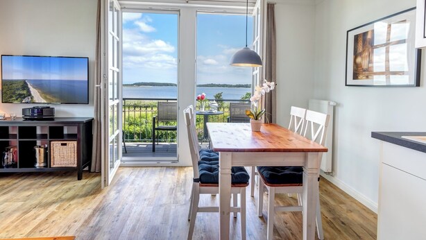A modern living room with a wooden floor, a TV on a black stand, a dining table with chairs, a pendant light, and a balcony with a view of the water.