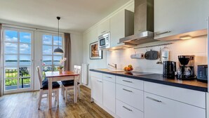 A modern kitchen with white cabinets, a black countertop, and stainless steel appliances. There is a dining area with a wooden table and chairs, and a view of the outdoors through large windows.