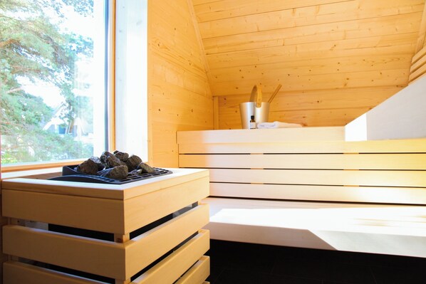 Interior de madera con sauna integrada, ventana con vista a follaje y una pequeña mesa con un recipiente metálico y utensilios.