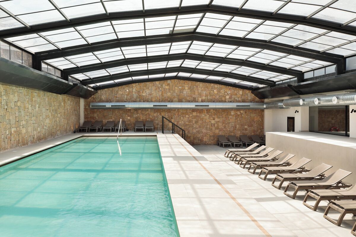 Una piscina interior con techo de vidrio, paredes de piedra y sillones de descanso a su borde.