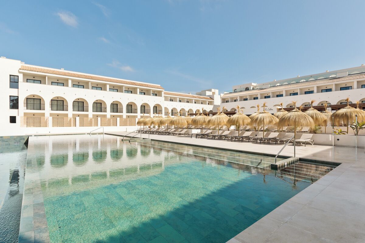 Ein Schwimmbecken mit Liegestühlen und Sonnenschirmen, ein Gebäude mit Rundbogenfenstern und ein klarer Himmel.