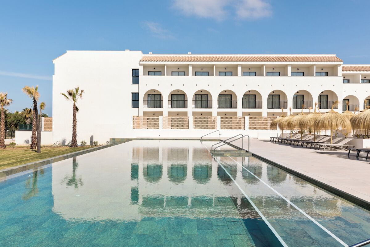 Un área de piscina con agua cristalina, un edificio blanco con ventanas arqueadas y palmeras.
