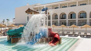 Parque acuático con toboganes y zona de piscina, rodeado por un edificio con balcones y sombrillas.