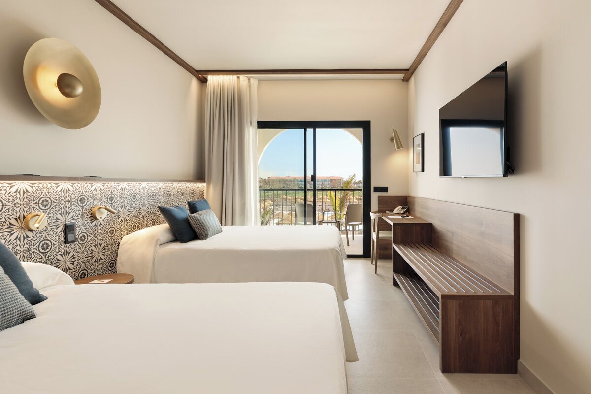 A hotel room with two beds, a TV, a desk, and a balcony with a view.