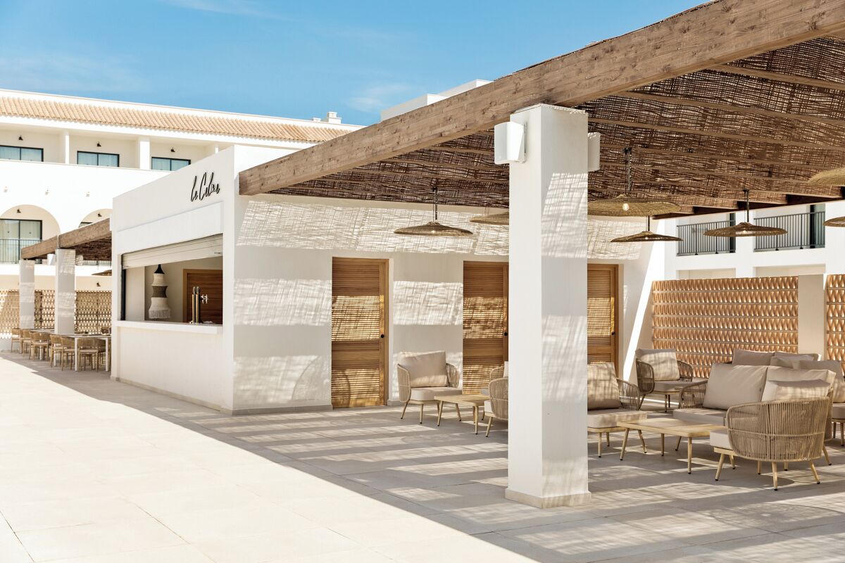 A modern outdoor seating area with wicker furniture, a wooden pergola, and a building with a white facade and arched windows.