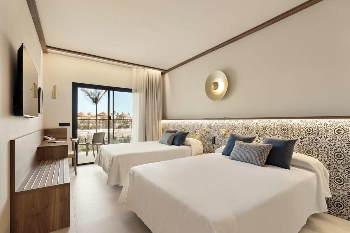 A hotel room with two beds, a desk, a chair, a TV, and a view of the outdoors.