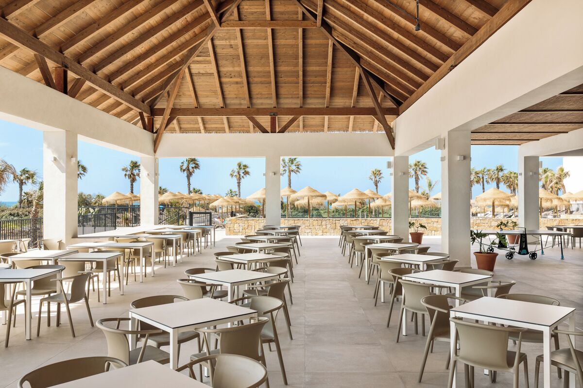 Un área de comedor al aire libre con mesas y sillas, techo de madera y vista a palmeras y sombrillas.
