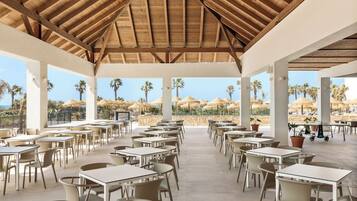 Un área de comedor al aire libre con mesas y sillas, techo de madera y vista a palmeras y sombrillas.