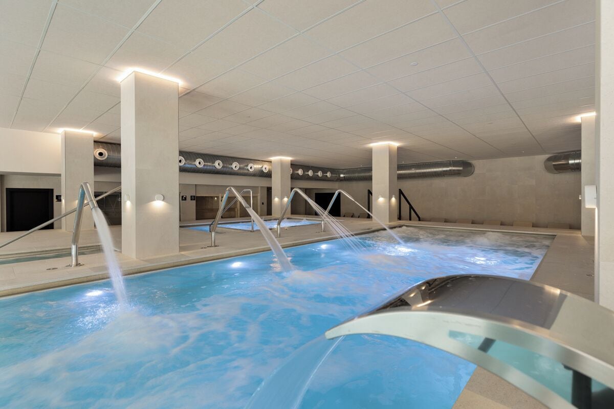Ein Innenpool mit mehreren Bahnen, einer zentralen Wasserspende und einem modernen Design mit freiliegenden Balken und Säulen.