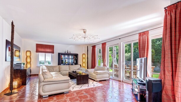 A spacious living room with a large sectional sofa, a coffee table, and a television. The room has a tiled floor and large windows with red curtains.