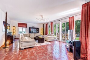 A spacious living room with a large sectional sofa, a coffee table, and a television. The room has a tiled floor and large windows with red curtains. - Vacation home Ahlbeck with sauna, wood-burning stove and space for up to 8 people (Ahlbeck)