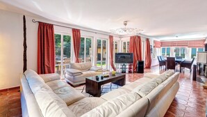 A spacious living room with a large sectional sofa, a coffee table, and a television. There are large windows with red curtains, and a dining area with a table and chairs is visible in the background.