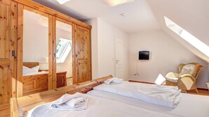 A bedroom with a wooden wardrobe, a bed with white linens, a wicker chair, and a TV on the wall.