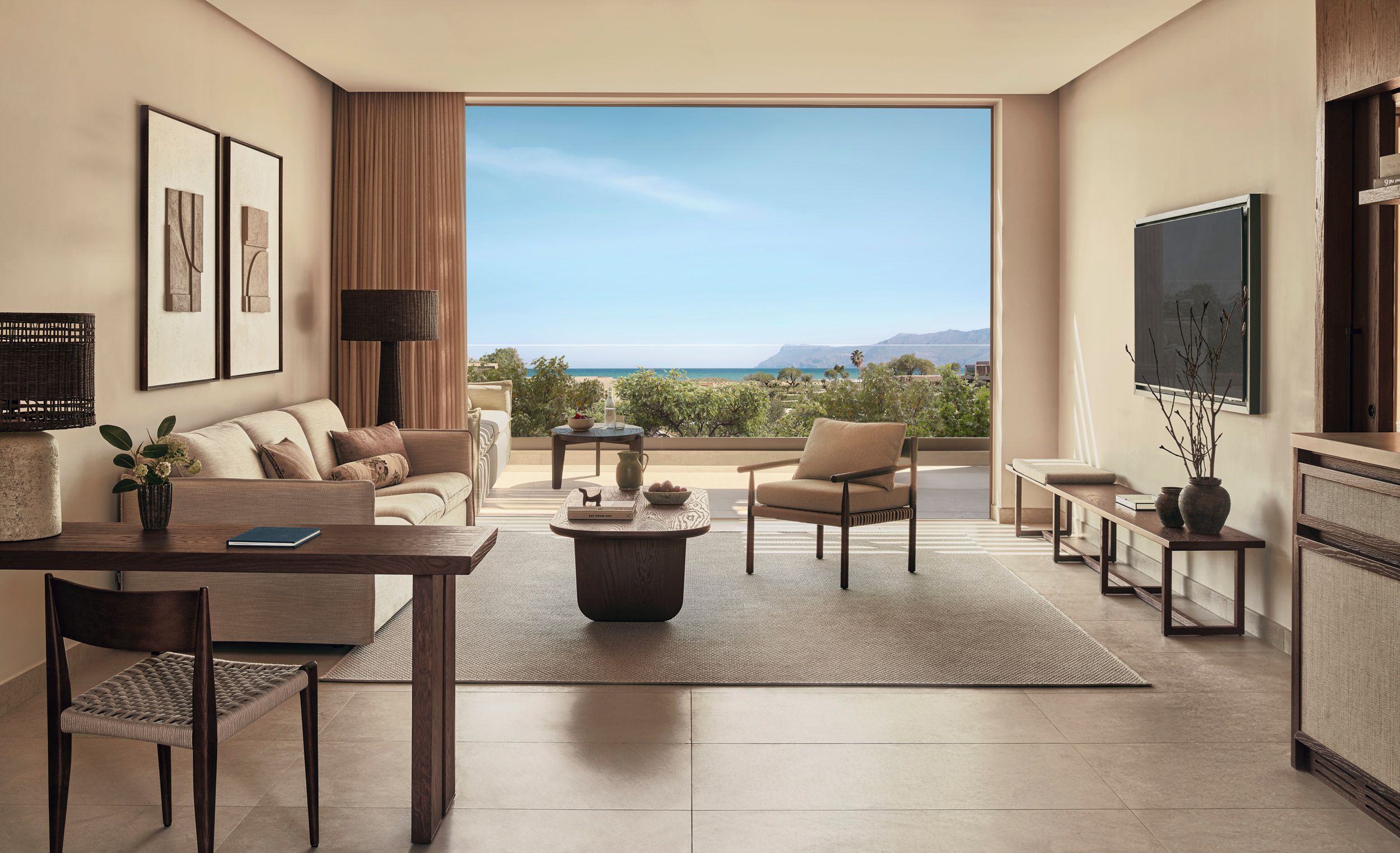 A modern living room with a sofa, armchairs, a coffee table, and a TV. The room has a large window with a view of the beach and mountains.