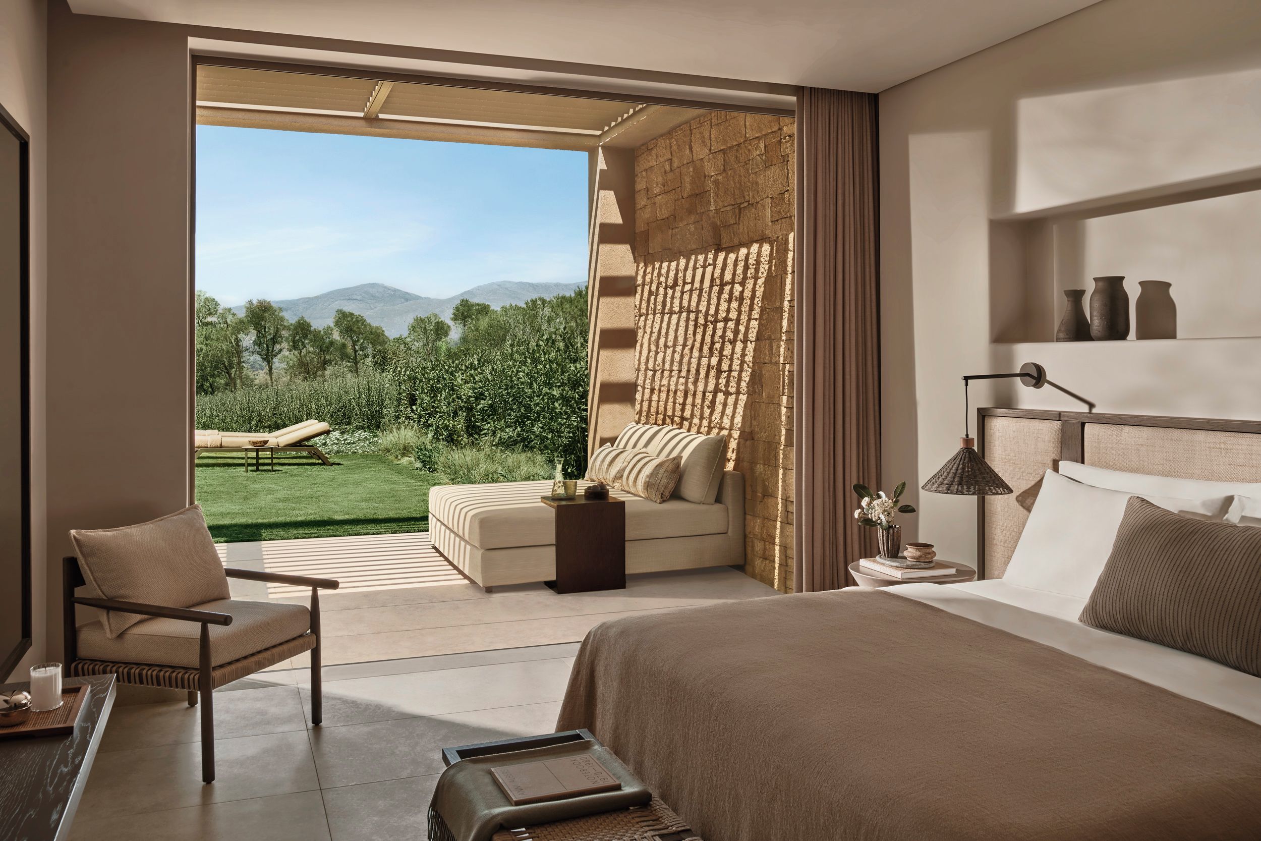 A bedroom with a large bed, a sofa, a chair, and a view of a garden and mountains.