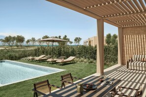 Uma área à beira da piscina com espreguiçadeiras, uma mesa com jarra e petiscos, e um pergolado com guarda-sol.