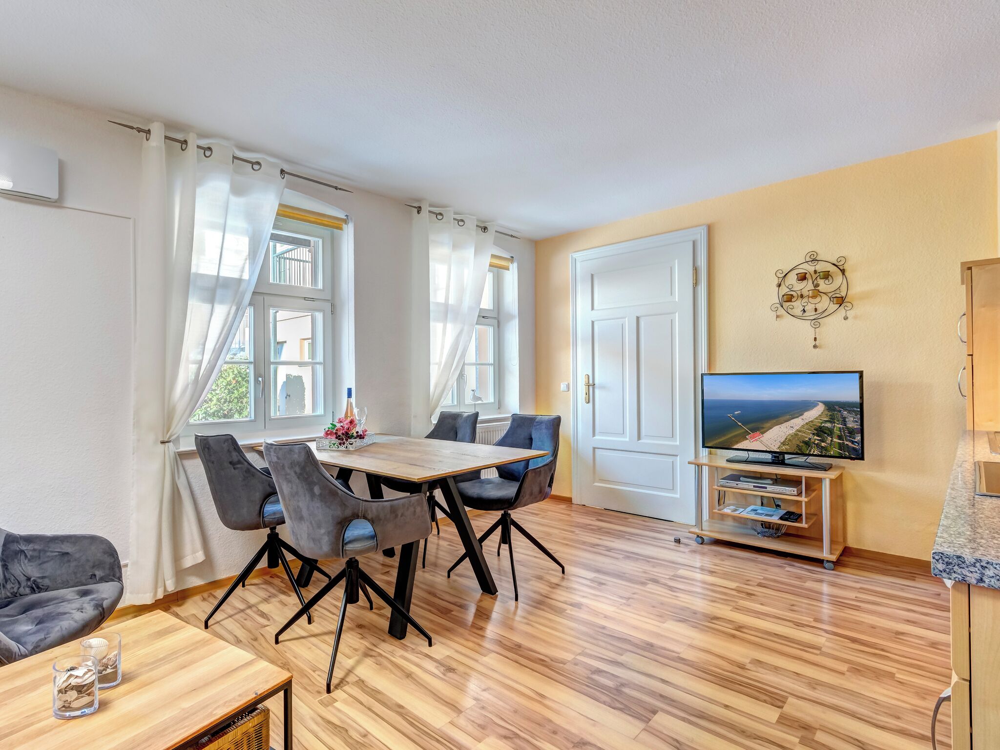 Ein modernes Wohnzimmer mit Holzboden, einem Esstisch mit Stühlen, einer Couch, einem Fernseher auf einem Schrank und einem Wandbild.