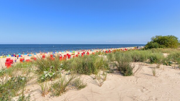A beach with numerous colorful umbrellas and beach chairs, sandy dunes, and a clear blue sky.