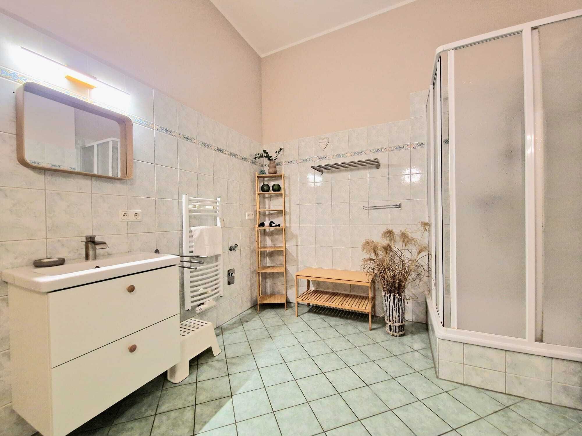 A bathroom with a white vanity, a mirror, a towel rack, a shelf with toiletries, a bench, and a shower area.