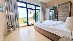 A modern hotel room with a large bed, a bedside table with a lamp, a large mirror, and a view of greenery outside.