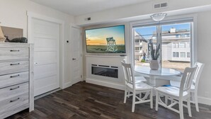 A dining area with a white table and chairs, a painting above the fireplace, and a view of the outside through large windows. - 1BR Beach Retreat - Pool, Parking & Boardwalk (Ocean City)