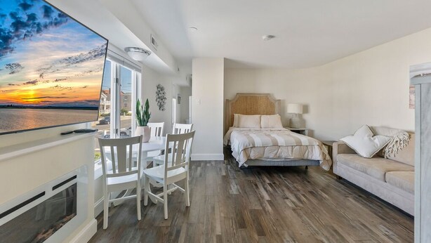 A modern bedroom with a large window, a bed, a bedside table, a chair, and a TV. - 1BR Beach Retreat - Pool, Parking & Boardwalk (Ocean City)