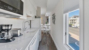 A narrow kitchen with a coffee maker, toaster, and sink. There is a window with a view of a street and buildings. - 1BR Beach Retreat - Pool, Parking & Boardwalk (Ocean City)