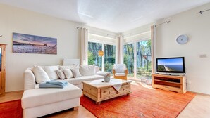 A modern living room with a sofa, a coffee table, a TV on a wooden stand, and a large window with a view of trees. - House Berlin, apartment Loddiner Höft in Loddin - in the best location near the Achterwasser (Loddin)