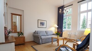 Uma sala de estar com um sofá listrado, uma mesinha com abajur, uma cadeira, um espelho e uma janela com cortinas.