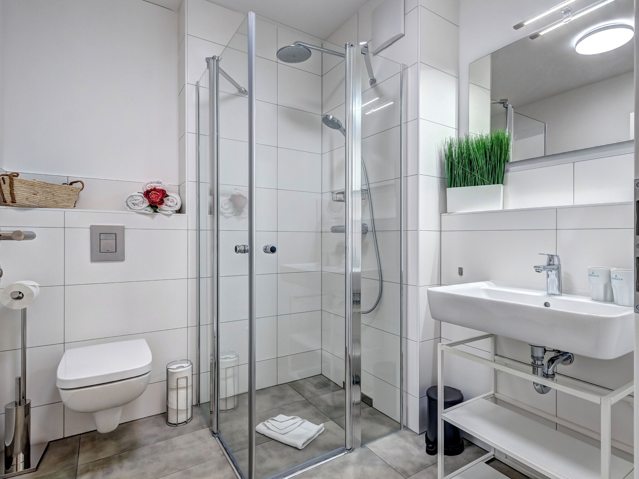 Une salle de bain moderne avec une douche à parois de verre, des toilettes blanches, un lavabo avec un miroir et une étagère contenant des articles de toilette.