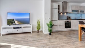 A modern living room with a flat-screen TV, wooden flooring, a white entertainment unit, and a kitchen with grey cabinets and a built-in oven. - Bernsteinquartier, vacation home 06b "Lachmöwe" in Kölpinsee - Baltic Sea vacation deluxe (Kölpinsee)