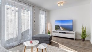 A modern living room with a large flat-screen TV, a grey sofa, two small white tables, and a white cabinet. - Bernsteinquartier, vacation home 06b "Lachmöwe" in Kölpinsee - Baltic Sea vacation deluxe (Kölpinsee)