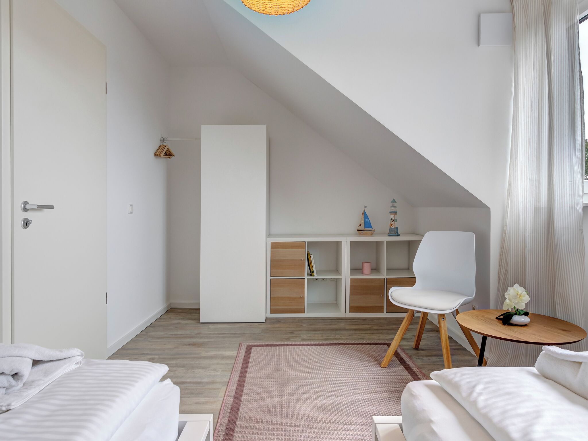 A modern, well-lit room with a sloped ceiling, white walls, and a wooden floor. It includes a bed, a white chair, a small round table with a vase of flowers, and a bookshelf with decorative items.