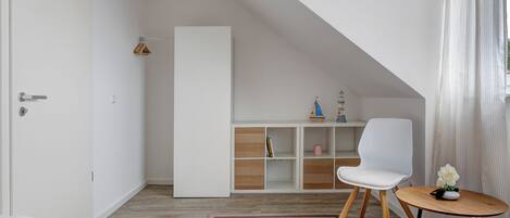 A modern, well-lit room with a sloped ceiling, white walls, and a wooden floor. It includes a bed, a white chair, a small round table with a vase of flowers, and a bookshelf with decorative items.
