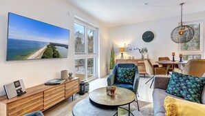 A modern living room with a large TV, comfortable seating, and a view of the sea.