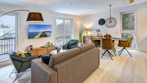 A spacious living room with a large flat-screen TV, a sofa, a dining table with chairs, and a balcony with a view of the city.