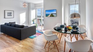 A modern living room with a black sofa, a glass dining table with white chairs, and a large window with a view of the sea.