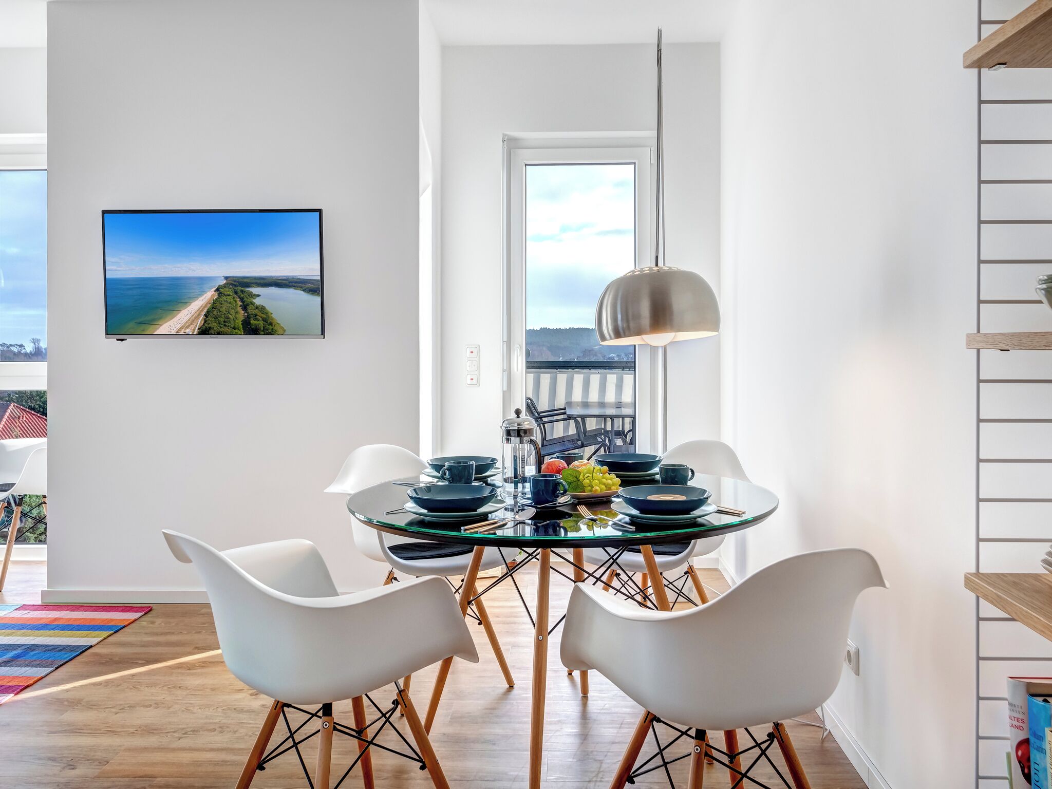 Et moderne spiseområde med et rundt bord, hvide stole og en hængende lampe. Et stort vindue giver udsigt til en strand og bybillede.