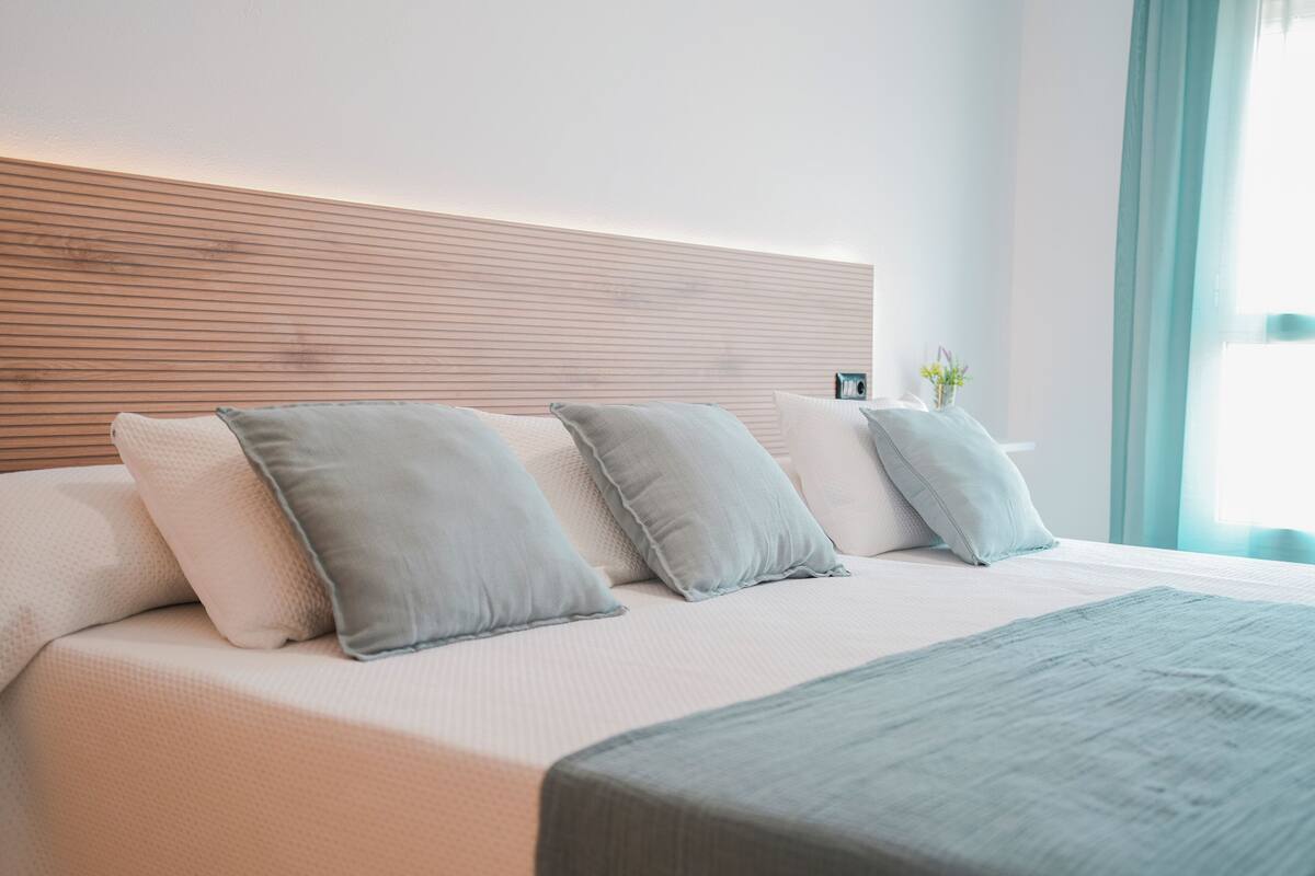 Een net opgemaakt bed met een houten hoofdbord, meerdere kussens en een nachtkastje met een kleine vaas.