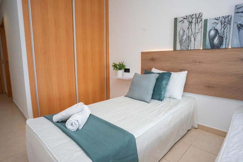 A neatly made bed with a green throw blanket and white towels, a wooden headboard, and a wall-mounted shelf with a plant.