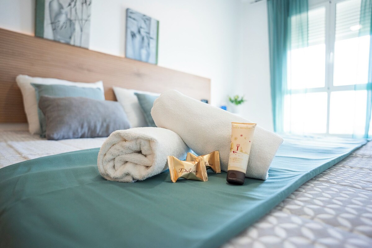 Cama bem arrumada com um edredom verde, uma toalha branca enrolada, um par de chinelos e um tubo de creme sobre a cama.