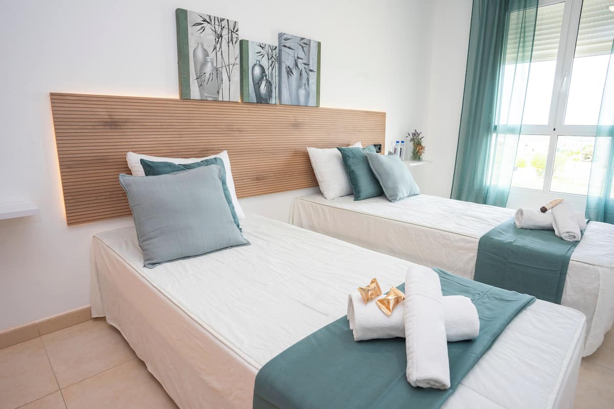 Een hotelkamer met twee bedden, elk met een houten hoofdeinde, witte beddengoed en blauwgroene accenten. Er liggen handdoeken en een klein vaasje op de bedden.